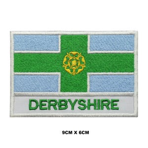 May include: Embroidered patch featuring the flag of Derbyshire, a white background with a green cross and a yellow floral emblem in the centre. The text "DERBYSHIRE" is written in white below the flag. The patch measures 9 cm by 6 cm.