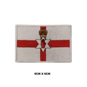 May include: An embroidered patch featuring the flag of Northern Ireland. The patch is white with a red cross and a red hand inside a six-pointed star. A gold crown sits above the star. The patch measures 9 cm by 6 cm.