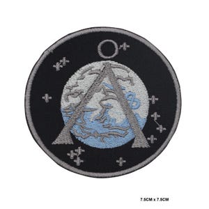 May include: A black and gray embroidered patch with a stylized image of a planet with a ring around it. The planet is surrounded by a circle of stars. The patch is 7.5 inches by 7.5 inches.