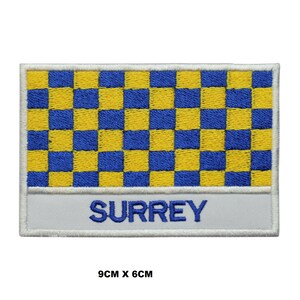 Pode incluir: Um remendo bordado branco com um padrão xadrez azul e amarelo e a palavra "SURREY" em letras brancas. O remendo mede 9 cm por 6 cm.