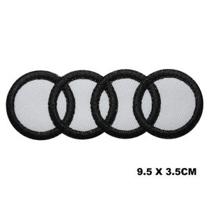 Può includere: Una toppa ricamata con quattro cerchi intrecciati con centri bianchi e bordi neri spessi. Il design è disposto orizzontalmente in una fila. Il testo 9.5 X 3.5CM è stampato in nero nell'angolo in basso a destra dell'immagine.