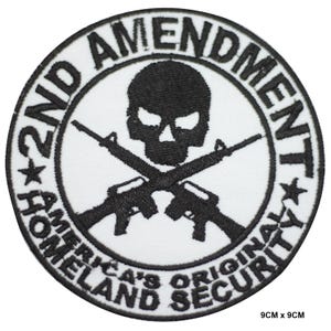 Könnte beinhalten: Ein weißer gestickter Aufnäher mit einem schwarzen Totenkopf und gekreuzten Knochen, zwei Gewehren und dem Text "2nd Amendment America's Original Homeland Security" in einem Kreis.