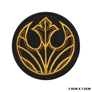May include: Circular black embroidered patch with a gold emblem. The emblem has a stylized design with curved lines and a central element. The patch measures 7.5cm x 7.5cm.