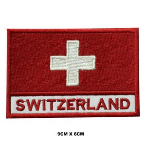 May include: Embroidered patch featuring the Swiss flag with a white cross on a red background. The patch also includes the word "SWITZERLAND" in white lettering on a red background. The patch measures 9 cm by 6 cm.