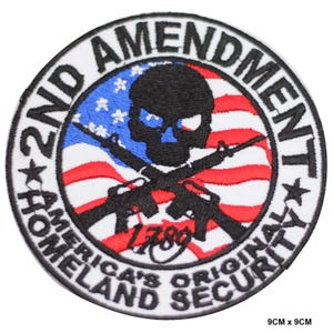 May include: A white, round patch with black text that reads "2nd Amendment America's Original Homeland Security" and a black skull with crossed rifles in front of an American flag with red and white stripes and blue stars. The year "1789" is written below the skull.