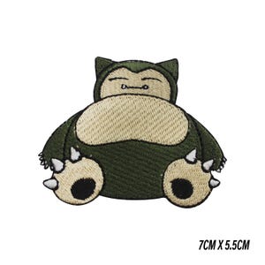 Peut inclure: Un écusson brodé représentant une créature vert foncé et beige aux yeux fermés, avec un gros ventre et des griffes blanches sur les pieds. L'écusson mesure 7 cm sur 5,5 cm et montre un personnage au corps arrondi, aux oreilles pointues et aux pieds sombres.