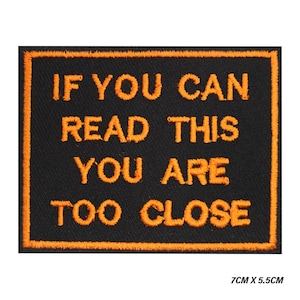 Może przedstawiać: Czarna prostokątna naszywka z pomarańczowym haftowanym tekstem: „IF YOU CAN READ THIS YOU ARE TOO CLOSE”. Naszywka ma pomarańczową obwódkę i wymiary 7 cm x 5,5 cm.