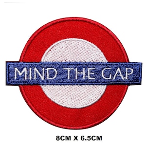 May include: Embroidered patch with the text "MIND THE GAP" in white on a blue banner, set within a red circle with a white centre. The patch measures 8cm x 6.5cm.