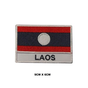 May include: Embroidered patch featuring the flag of Laos. The flag is red, blue, and white with a white circle in the center. The patch is 9 inches by 6 inches and has the word "LAOS" written below the flag.