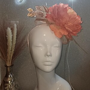 May include: A peach-coloured fabric flower with gold accents is attached to a white headband. The headband is on a white mannequin head.