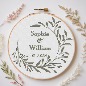 Peut inclure: Un cerceau de broderie blanc avec un motif de point de croix représentant une couronne de feuilles avec les noms "Sophia & William" et la date "24.6.2024" au centre.