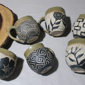 Puede incluir: Seis tazas de cerámica con diseños en blanco y negro sobre un fondo verde claro. Los diseños incluyen patrones geométricos, flores y hojas.