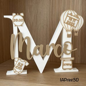 Peut inclure: Une grande lettre M blanche avec le prénom Marco en écriture dorée. Les décorations dorées incluent un réveil indiquant 20:32, deux montgolfières avec la date 19.11.2025, un body bébé marqué 52 cm et une balance affichant 3765g.
