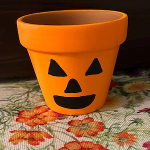 May include: Orange terracotta planter with a black painted jack-o'-lantern face. The planter is sitting on a floral fabric.