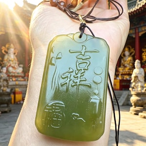May include: A rectangular olive green jade pendant featuring intricate relief carvings of Chinese characters and a lantern motif. The smooth, polished stone is suspended from a dark brown cord with small wooden beads, showcasing a translucent quality.