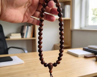 May include: A string of dark brown wooden beads arranged in a circular loop. The beads are uniform in size and shape, featuring a single larger, intricately carved bead at the bottom, followed by a short cord ending in four smaller beads.