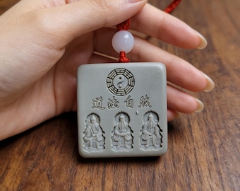 Puede incluir: Un colgante cuadrado gris con un cordón rojo. El colgante presenta un símbolo de Yin Yang, caracteres chinos y tres figuras talladas. El colgante está sujeto por una mano.