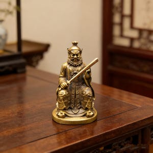 May include: A small, detailed brass statue of a seated figure holding a sword. The figure has a long beard and is wearing ornate robes. The statue is on a round base and sits on a dark wooden surface.