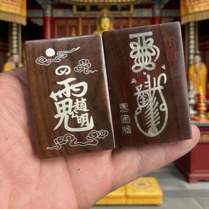 May include: Two rectangular wooden tiles with silver inlay. One tile features Japanese characters and cloud designs, the other has more characters and a fish-like design. The tiles are a dark brown color.