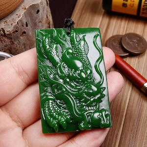 Puede incluir: Colgante rectangular verde jade con un detallado grabado de un dragón. La cabeza y el cuerpo del dragón están intrincadamente esculpidos, con un cordón negro adjunto en la parte superior. El colgante se sostiene en una mano, con una superficie de madera en el fondo.