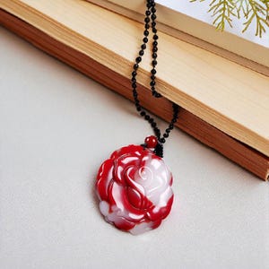 Puede incluir: Un collar con colgante de rosa roja y blanca. El colgante es un diseño de rosa tallada con colores rojo y blanco. El collar tiene una cadena de cuentas negras. El colgante está sobre una superficie gris claro junto a un libro de madera.