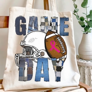 May include: Cream tote bag with a football theme. It features a white helmet, a brown football with a pink ribbon, and the words "GAME DAY" in denim patchwork. The bag is made of canvas.