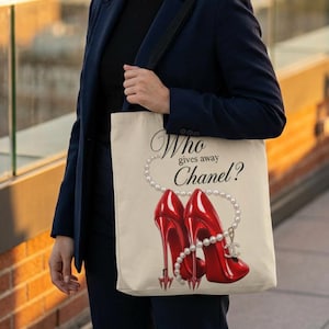 Könnte beinhalten: Cremefarbene Tragetasche mit einem Design aus zwei roten High Heels, einer Perlenkette und dem Text "Who gives away Chanel?". Die Tasche hat einen schwarzen Riemen und wird von einer Person getragen.