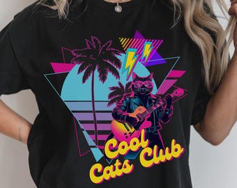 May include: Black t-shirt featuring a retro design with a cat playing a guitar, wearing sunglasses and a leather jacket. The design includes palm trees, lightning bolts, and the text "Cool Cats Club" in yellow and pink.