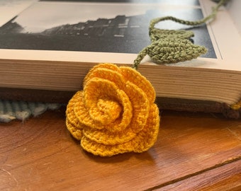 Op de afbeelding: Een gehaakte gele roos boekenlegger met groene bladeren. De boekenlegger ligt op een boek en een houten oppervlak.