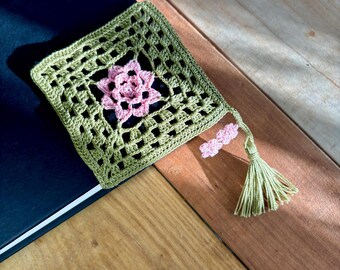 Op de afbeelding: Een groen gecrochet vierkant met een roze bloem in het midden. Het vierkant heeft een groene kwast aan een hoek.