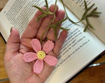 Op de afbeelding: Een handgemaakte gehaakte bladwijzer met een roze bloem en een gele kern, plus groene bladeren. De bladwijzer ligt op een open boek en benadrukt het delicate bloemendesign en het vakmanschap. Voor boekenliefhebbers.