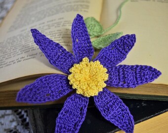Puede incluir: Un marcapáginas de ganchillo morado con un centro amarillo, descansando sobre un libro cerrado. El marcapáginas tiene forma de flor con cinco pétalos y un tallo con dos hojas verdes. El libro está cerrado y tiene una cubierta negra.