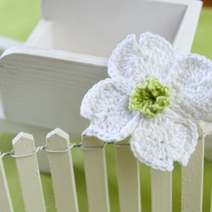 Peut inclure: Gros plan sur une fleur blanche au crochet avec un centre vert, posée sur une clôture blanche. Une petite brouette en bois blanche est en arrière-plan, sur un fond vert clair. La fleur est le point central.