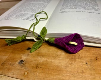 Op de afbeelding: Een gehaakt bladwijzer met een groen blad en een paarse bloem. De bladwijzer ligt op een boek.