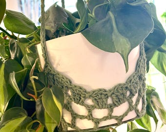 Op de afbeelding: Een groene gehaakte plantenhanger met een witte keramische pot en een groene, bladerrijke plant die eraan hangt.