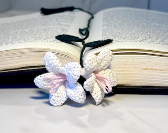 Op de afbeelding: Een witte gehaakte bloemenbladwijzer met roze accenten en groene gehaakte stelen. De bladwijzer ligt op een gesloten boek.