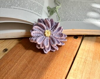 Op de afbeelding: Een gehaakte paarse bloemenbladwijzer met een geel centrum, liggend op een houten oppervlak. De bladwijzer is bevestigd aan een groene gehaakte ketting.