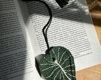 Op de afbeelding: Een gehaakt groen blad met witte aderen. Het blad hangt aan een boek met tekst over een spookhuis.