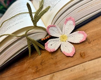 Op de afbeelding: Een gehaakt bladwijzer met een roze en witte bloem en groene bladeren. De bladwijzer ligt op een boek.