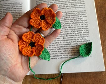 Op de afbeelding: Een handgemaakt gehaakt bladwijzer met twee oranje bloemen en groene bladeren. De bladwijzer ligt op een boek met tekst over een spookhuis.
