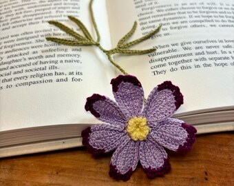 Op de afbeelding: Een gehaakte paarse bloemenbladwijzer met een gele kern en een groene stengel. De bladwijzer ligt op een open boek.