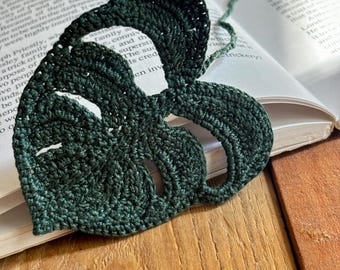 Op de afbeelding: Een donkergroene, hartvormige gehaakte bladwijzer met een bladachtig ontwerp, rustend op een open boek. De bladwijzer is gemaakt van garen en heeft een getextureerd uiterlijk. Een groene garenstreng loopt vanaf de bovenkant van de bladwijzer.