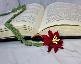 Op de afbeelding: Een groene gehaakte bladwijzer met een rode en gele gehaakte bloem eraan bevestigd. De bladwijzer ligt op een gesloten boek.