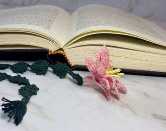 Op de afbeelding: Een roze gehaakte bloemenbladwijzer met gele accenten, bevestigd aan een groene gehaakte ketting met een kwast.