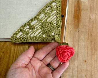 Puede incluir: Marcapáginas de ganchillo verde con una rosa de ganchillo rosa unida. El marcapáginas tiene forma de triángulo y presenta un delicado patrón de encaje.