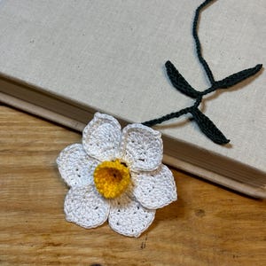 May include: A white crocheted daffodil bookmark with a yellow center and green crocheted stem.