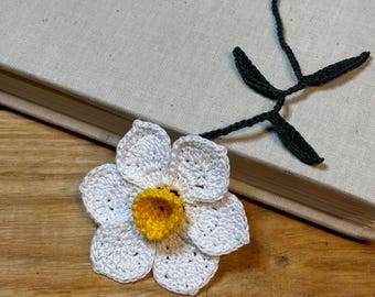 Op de afbeelding: Een witte gehaakte narcissen-bladwijzer met een geel centrum en een groene gehaakte stengel.