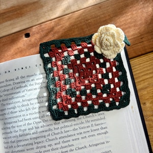 May include: A handmade crochet bookmark in a granny square pattern, featuring a cream-colored flower accent. The bookmark has a dark green border, with red, white, and dark green squares. It is placed on a book.