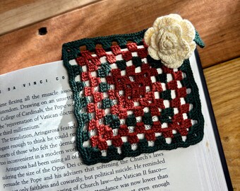 Op de afbeelding: Een handgemaakte gehaakte bladwijzer in een granny square patroon, met een crèmekleurig bloemaccent. De bladwijzer heeft een donkergroene rand, met rode, witte en donkergroene vierkanten. Het ligt op een boek.