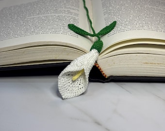 Op de afbeelding: Een wit gehaakt calla lelie boekenlegger met groene gehaakte bladeren en een geel gehaakt midden. De boekenlegger is geplaatst in een open boek.